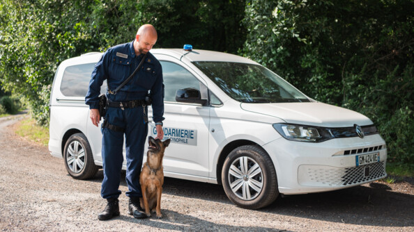 Un maître de chien de plein-pied, en combinaison bleue-marine, avec un malinois assis à ses pieds qui le regarde. Au fond le véhicule blanc de l'équipe