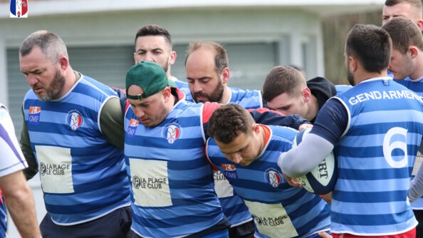 La premi&egrave;re et deuxi&egrave;me ligne d'une &eacute;quipe de rugby, au maillot bleu, se pr&eacute;pare &agrave; la mếl&eacute;e. A droite, un joueur de dos tient le ballon sous son bras gauche