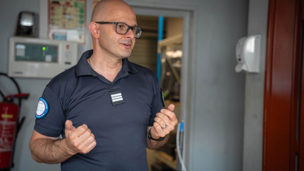 Un homme d'âge moyen se tient debout, face à l'objectif, dans une pièce, derrière un comptoir. Il discute, s'animant avec les mains. Il porte un t-shirt bleu marine et des lunettes noires. L'homme est chauve.