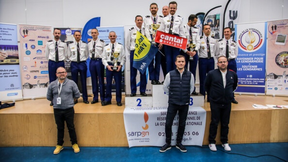 11 coureurs en tenue chemise blanche avec leurs médailles au cou sur une scène et le podium; en fond des kakémonos gendarmerie et sponsors; au pied de la scène trois hommes en civils mains derrière le dos