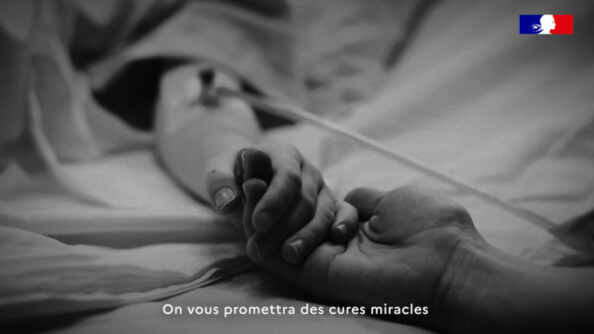Image en noir et blanc : La main d'une personne tient celle d'une autre personne, allongée dans un lit, recouverte d'un drap, avec un cathéter au creux du bras. On n'aperçoit que les mains et les avant-bras de deux personnes.