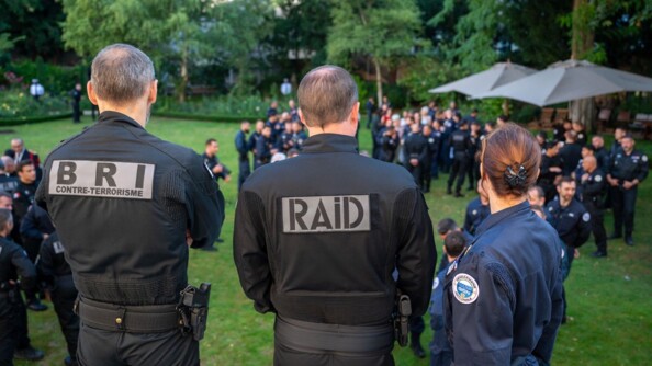 Trois militaires de la BRI, du RAID et du GIGN se tiennent debout, de dos, face à un grand jardin, dans lequel de nombreuses personnes discutent. Leur unité d'appartenance est reconnaissable par l'insigne qu'ils portent sur leur veste.