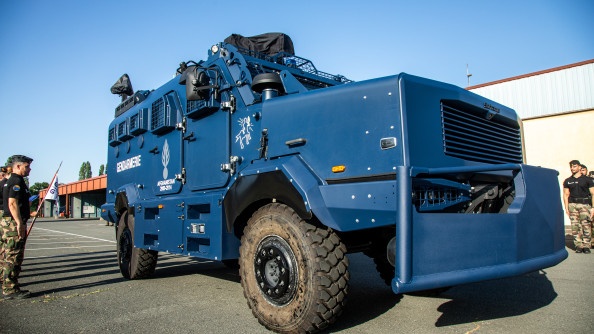 La photo représente un véhicule blindé de la gendarmerie avançant devant des mililtaires alignés au bord de la route