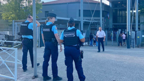 Trois gendarmes de dos dont une de profil et souriante devant les abords d'une école