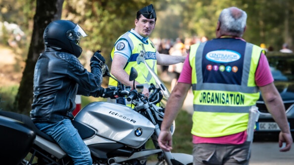 Un motard, de profil droit, sur une moto grise discute avec un gendarme de trois quart avant droite, revêtu d'une chasuble jaune, qui le regarde en montrant une direction de son bras gauche. Devant le motard, de dos, un homme portant une chasuble jaune avec la mention "organisation"