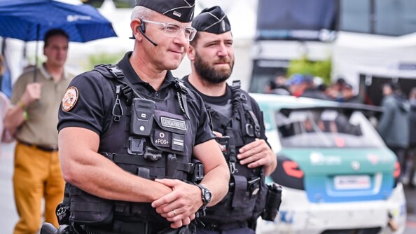 Deux gendarmes en noir souriants. Derrière eux flou, à droite, une voiture de course et à gauche un homme portant un parapluie