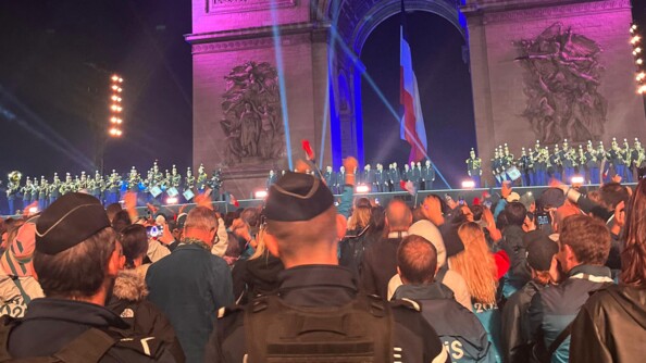 Sur une grande sc&egrave;ne install&eacute;e au pied de l'Arc de Triomphe, o&ugrave; flotte le drapeau fran&ccedil;ais, les musiciens de la Garde R&eacute;publicaine occupent les deux tiers de la sc&egrave;ne ; &agrave; droite, v&ecirc;tus de blanc, les athl&egrave;tes olympiques et paralympiques saluent la foule amass&eacute;e devant la sc&egrave;ne. Au premier plan, deux gendarmes d&eacute;partementaux, de dos, issus d'une des compagnies de marche mobilis&eacute;es pour assurer la s&eacute;curisation des festivit&eacute;s.