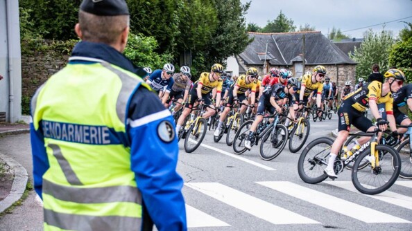 Gendarme vu de dos, avec une chasuble fluo, au bord de la route, sécurisant le passage d'un peloton de cyclistes à travers un village.
