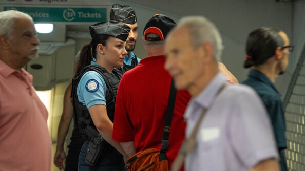 Deux gendarmes patrouillent dans une station de métro souterraine. Autour d'eux évoluent des passagers. Un panneau situé derrière eux indique les directions de Porte de Clignancourt et de Bagneux, situées sur la ligne numéro 4.