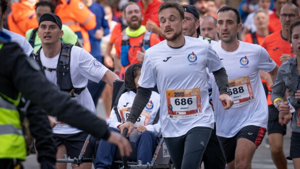 Une foule de coureurs, vêtus d'un T-shirt blanc avec un numéro sur le ventre, courent côte à côte. L'un d'eux pousse un fauteuil roulant.
