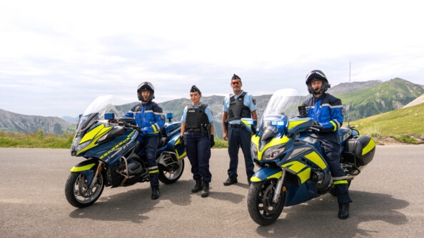Deux gendarmes motocyclistes, installés sur leur moto, à l'arrêt, posent face à l'objectif, sur une route de montagne. Entre eux, deux motards se tiennent debout sur la chaussée.