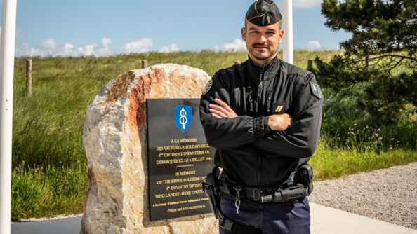 Un gendarme mobile pose devant un momument à la mémoire de la 8e division d'infanterie. au pied du monument, un casque d'époque de l'unité