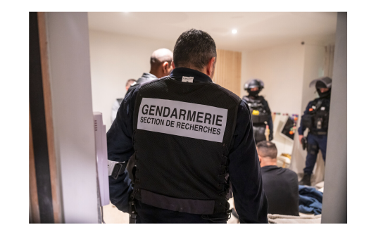 La photo représente un gendarme vêtu d'une tenue noire, portant dans le dos l'inscription " Gendarmerie - Section de recherches", qui se tient debout, dans une pièce, en présence d'autres personnes .