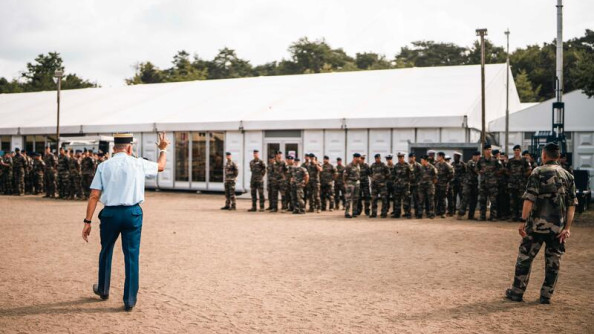 Un gendarme passe devant un groupe de militaires en tenue kakie, effectuant le signe de la victoire de sa main droite.