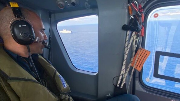 Vue sur la mer et le porte hélicoptères Mistral depuis le hublot d'un hélicoptère de la gendarmerie nationale. Au premier plan un militaire de l'équipage.