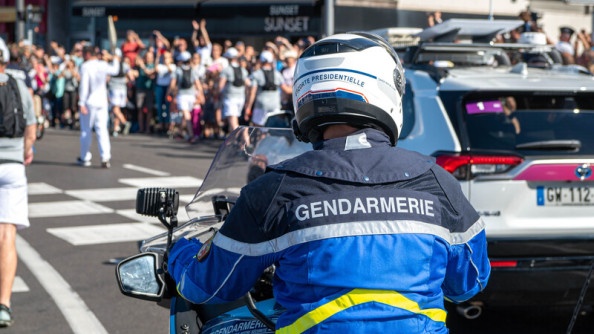 Un gendarme sur sa moto, que l'on voit de dos, évolue sur la route, au bord de laquelle se tient une foule très dense.