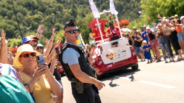 Un gendarme, sifflet à la bouche, sur une route, autour de laquelle la foule s'agglomère. sur la route des véhicules rouge avec des animations
