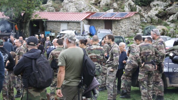 De nombreux militaires sont rassemblés sur une pelouse, à proximité d'une bergerie corse. On aperçoit un véhicule de la gendarmerie. Quelques civils se mêlent au groupe.