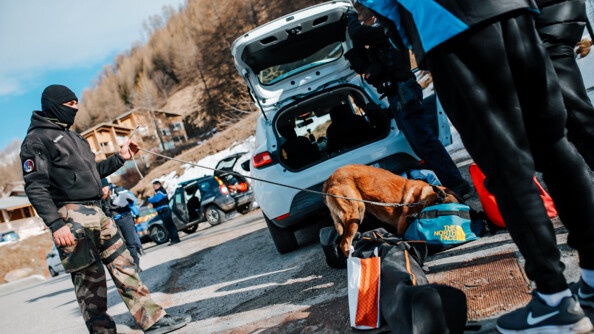 Un maître de chien tient par la laisse son malinois en train de fouiller dans des sacs au pied de l'arrière d'un véhicule. a droite, les jambes et pied d'une persone en surv^tement noir. A gauche au fond, des véhciules de gendarmerie et des gendarmes, en hauteur la montagne et des chalets