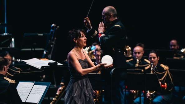 Une femme tenant un globe lumineux dans les mains avec une robe longue grise sur une scène au milieu de l'orchestre de la garde républicaine