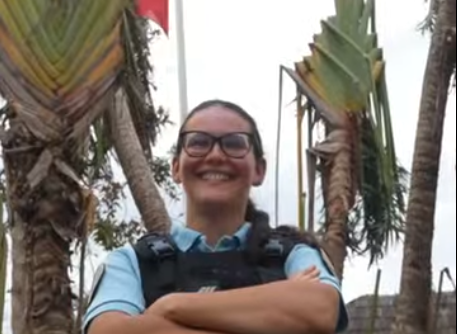 Femme gendarme souriante, avec des lunettes, les bras croisés sur la poitrine, debout, sous un palmier