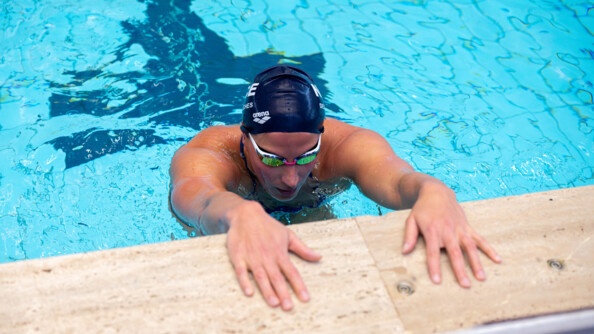 Une nageuse revêtant un bonnet bleu marine ainsi que des lunettes de nage, récupère, au bord du bassin, les mains posées sur le rebord de la piscine