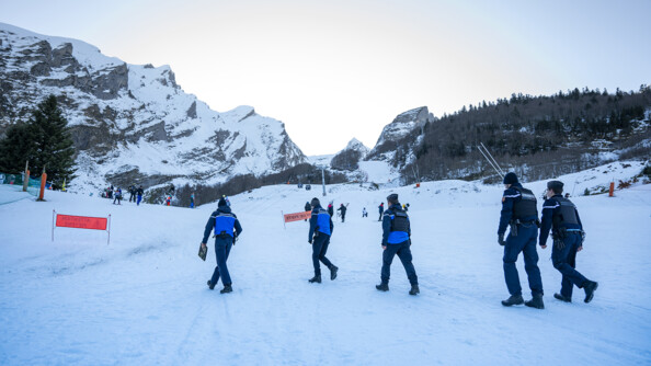 Cinq gendarmes en train de monter sur les pistes enneigées, en arrière-plan les montagnes et pistes avec des skieurs, à droite une pente d'arbres