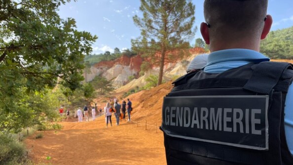 Un gendarme vu de dos, avec la mention GENDARMERIE sur son gilet pare-balles, est posté au bord d'un chemin couleur ocre, bordé de quelques arbres. Au loin, plusieurs presonnes marchent ou sont statiques.