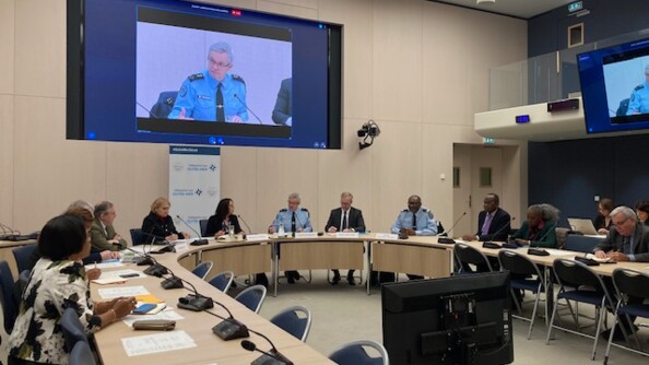 Dans une vaste salle aux murs beiges, des hommes et des femmes, civils et gendarmes en uniformes, sont assis autour d'une grande table circulaire, avec des micros devant chacun d'eux. Au mur est accroché un grand écran télé, sur lequel apparait un homme en uniforme.
