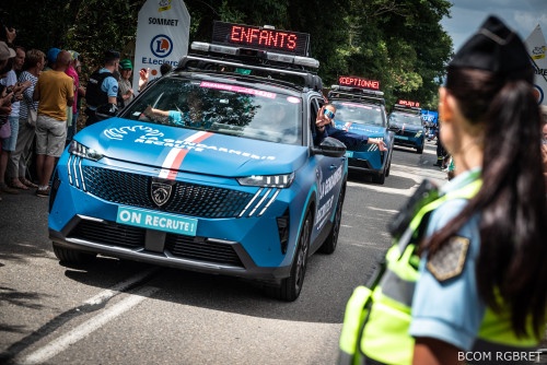 Une gendarme en uniforme, avec un gilet tactique jaune fluo et une queue de cheval, vue de trois-quart dos, est positionnée sur le bord de la route, tandis que les véhicules gendarmerie de la caravane du Tour de France passent devant elle. De l'autre côté de la chaussée sont massées de nombreuses personnes.