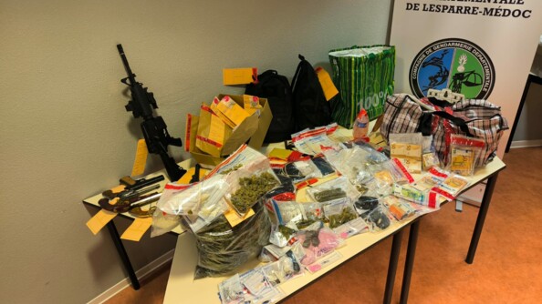 Sur une table beige sont posés de nombreux objets, recouvrant toute sa surface : billets de banque, sac transparents contenant des produits stupéfiants, armes, pochettes scellées, cabas... En second plan se dresse un kakémono portant la mention "Compagnie de gandarmerie départementale de Lesparre-Médoc