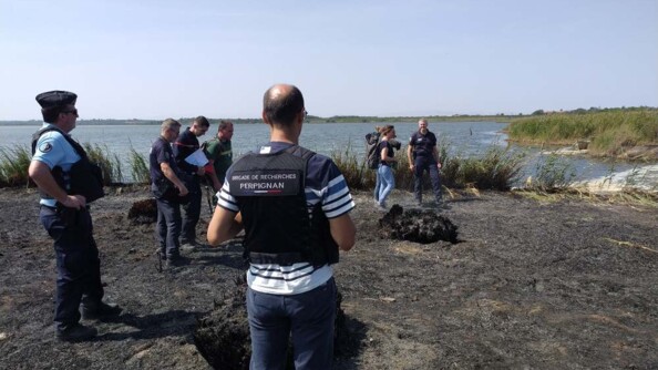 Sur un sol calciné, en bordure de mer, des gendarmes se tiennent debout, de dos. Dans leur dos est inscrite la mention "Brigade de recherches Perpignan". Deux personnes en tenue civile, ainsi qu'un sapeur-pompier, évoluent au milieu du groupe de gendarmes.