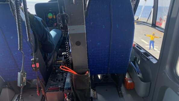 Photo de l'intérieur de l'hélicoptère de la gendarmerie nationale. Vue de dos sur le pilote et le co-pilote pendant la manoeuvre d'appontage. Par le hublot, à droite, vue sur le pont du porte hélicoptères et l'agent de piste qui guide l'équipage.