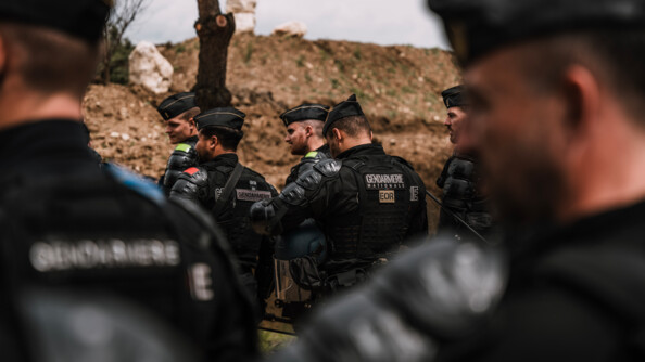 Un groupe de 5 gendarmes mobiles devant un talus de terre. En flou au premier plan, deux autres gendarmes mobiles qu'on devine