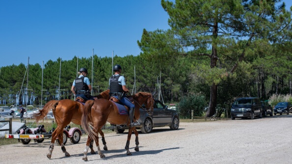 Deux gendarmes montés sont en train de patrouiller le long du lac de Biscarosse. le long de la voie, sur la gauche, des véhicules et une remorque de bateau sont stationnés. Au troisième plan, la pinède. Sur la gauche les bateaux amarrés dont on voit les mâts