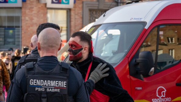 Photo de deux gendarmes, de dos, dans la rue, parlant avec un carnavaleux, au visage maquill&eacute; en noir et rouge, lors du carnaval de Bergues, devant un camion de pompiers
