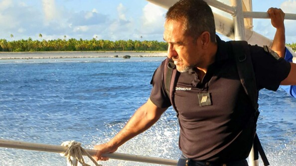 Sur un bateau, l'adjudant Teihotua se tient à une rembarde. Derrière lui, la mer et une île avec des palmiers.