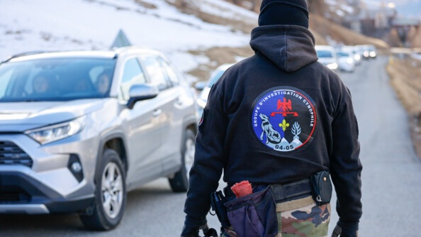 De dos un gendarme en treillis et haut noir du Groupe d'investigation cynophile devant une file de véhicules sur une route en bord de montagnes