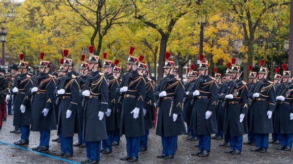 Sur un sol pavé, devant des arbres feuillus aux couleurs de l'automne, se tiennent des militaires de la Garde républicaine. Vêtus d'un long manteau noir, d'un pantalon bleu marine et du traditionnel chapeau noir à plumes rouges, ils tiennent leur fusil serré contre leur poitrine.