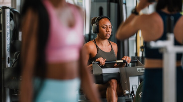 Une sportive vêtue d'une tenue grise, effectue un exercice sur un appareil de musculation. Elle porte un casque sur les oreilles.