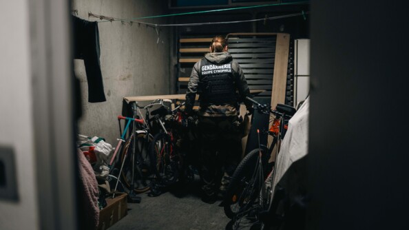 Une femme gendarme, portant la mention "Gendarmerie &eacute;quipe cynophile" dans le dos, fouille une cave sombre dans le sous-sol d'un immeuble, dans laquelle diff&eacute;rents objets sont entrepos&eacute;s, comme des v&eacute;los.