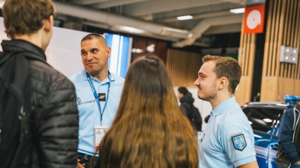 Deux gendarmes, souriants, vêtus d'un polo bleu ciel, discutent avce deux jeunes gens, qui se tiennent debout, tournant le dos à l'objectif. La scène se déroule au salon de l'Auto 2024, à Paris.