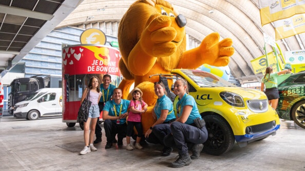 Qautre gendarmes posent accroupis aux côtés de deux fillettes, devant un véhicule jaune décapotable, dans lequel est installé un immense lion en peluche jaune.
