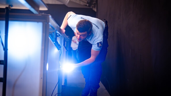 Dans un espace intérieur et sombre, un gendarme avanceles jambes fléchies et le corps plié au niveau de la taille. Il est vêtu d'un pantalon sombre et d'un polo bleu ciel. On aperçoit au niveau du dos l'inscription Gendarmerie. Il tient dans sa main gauche une lampe torche à l'aide de laquelle il inspecte le dessous d'un élément.