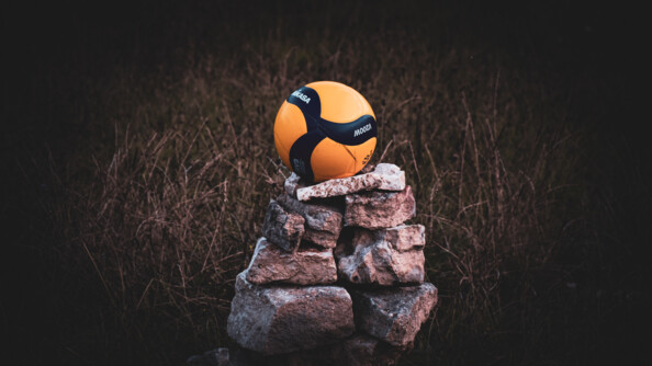 Au centre de l'image, un ballon de volley noir et jaune-orangé, posé sur un monticule de pierres, au milieu d'un champ.