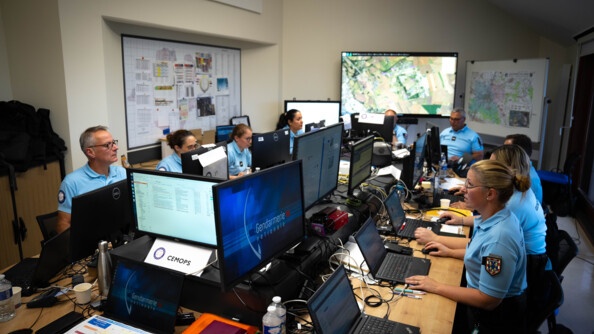 Neuf gendarmes, cinq femmes et quatre hommes, en polo bleu manches courtes sont assis à des bureaux qui se font face à face. Ils sont tous installés devant des ordinateurs. Au fond de la salle, se trouve un grand écran où est projetée une cartographie. Une autre carte est affichée sur un tableau à droite.  A gauche, sur le mur, un panneau où sont affichés des documents.