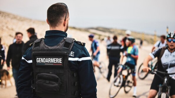 Au premier plan, un gendarme de dos, portant un gilet pare-balles siglé GENDARMERIE, par dessus une polaire. Au second plan, plusieurs cyclistes, pied à terre, et des gens à pied, un autre gendarme plus loin, dans un paysage minéral (Mont Ventoux), sous un ciel gris.