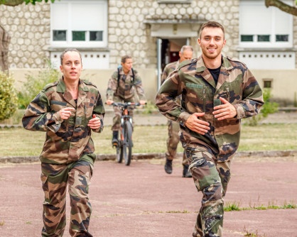Deux militaires ( une jeune femme à gauche et un jeune homme à droite) en tenue camouflage courent côté à côte.
