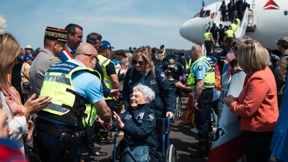 Une vétérante de la 2e guerre mondiale en fauteuil roulant serre la main d'un gendarme des transports aériens. Une foule de civils et militaires les entourent. Au fond à droite, la tête d'un avion