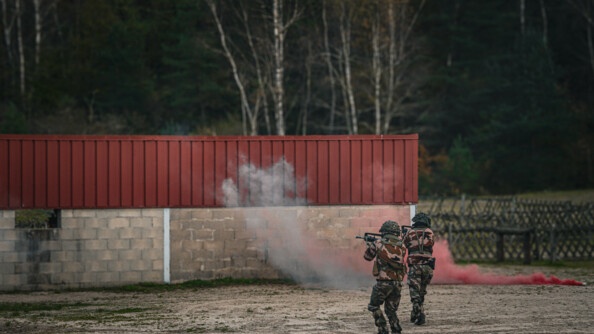 Deux militaires de dos, en treillis, casques sur la tête, progressent sur un terrain vague en pointant droit devant. Devant eux se dresse un mur de parpaings surmonté de tôle rouge.